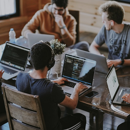 Four people sit around a table, each working on laptops. One wears headphones and types, while others are focused on their screens. There is a water bottle and a plant on the table. The setting appears casual and collaborative.