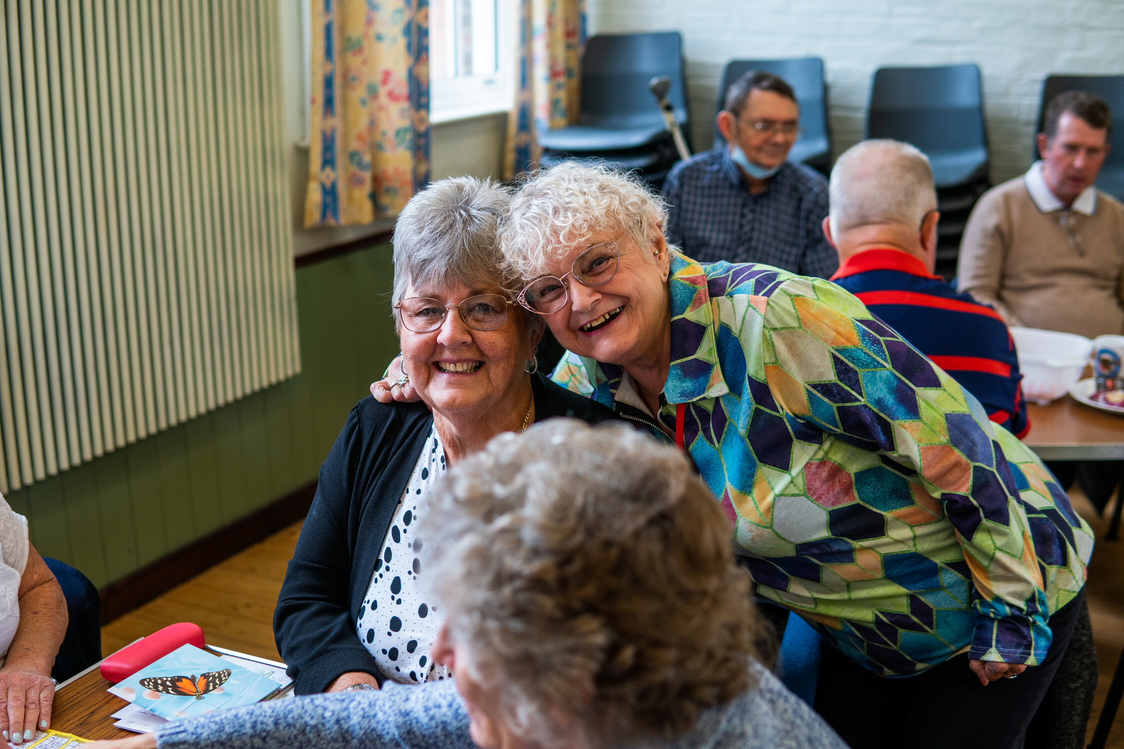 Two older women smile and pose together at a table in a community room, while others sit and talk in the background. The atmosphere is warm and friendly, with colorful decor on the walls and curtains.