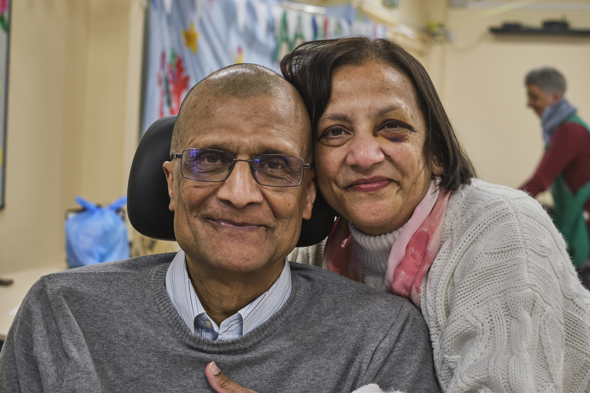 A man and woman sat together smiling in a community centre. The man is in a mobility chair.