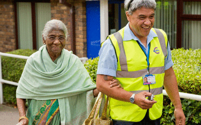Dial-A-Ride member of staff is walking alongside and older woman arm in arm.