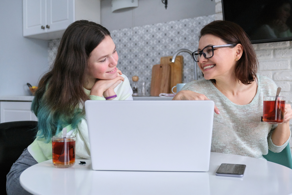 Mother and teenage daughter sitting at the kitchen table in front of a laptop. They are smiling.