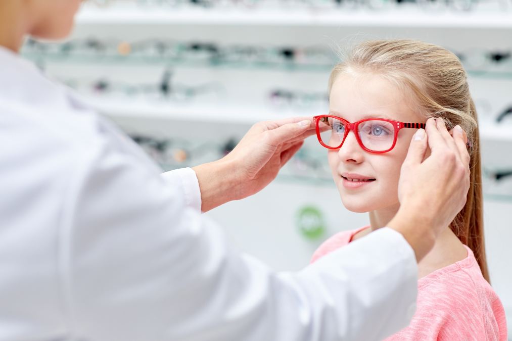 Blonde child getting red glasses fitted by an optician