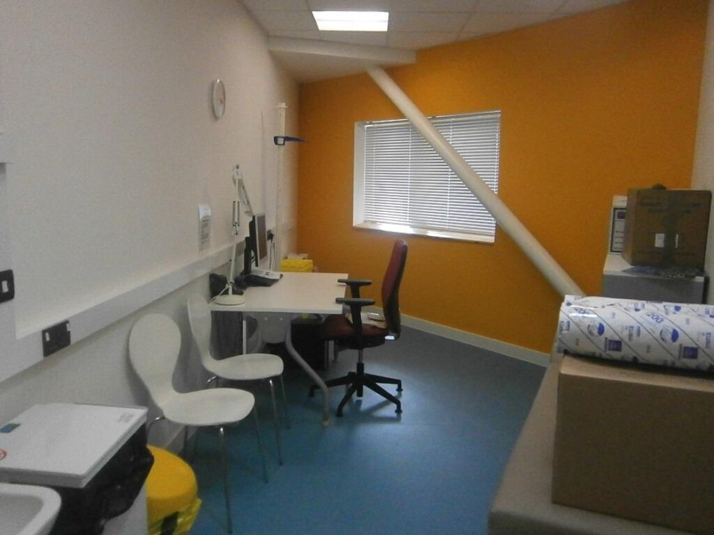 Small therapy room with orange wall, blue floor, desk with computer, white chairs, window with blinds, and visible medical supplies.