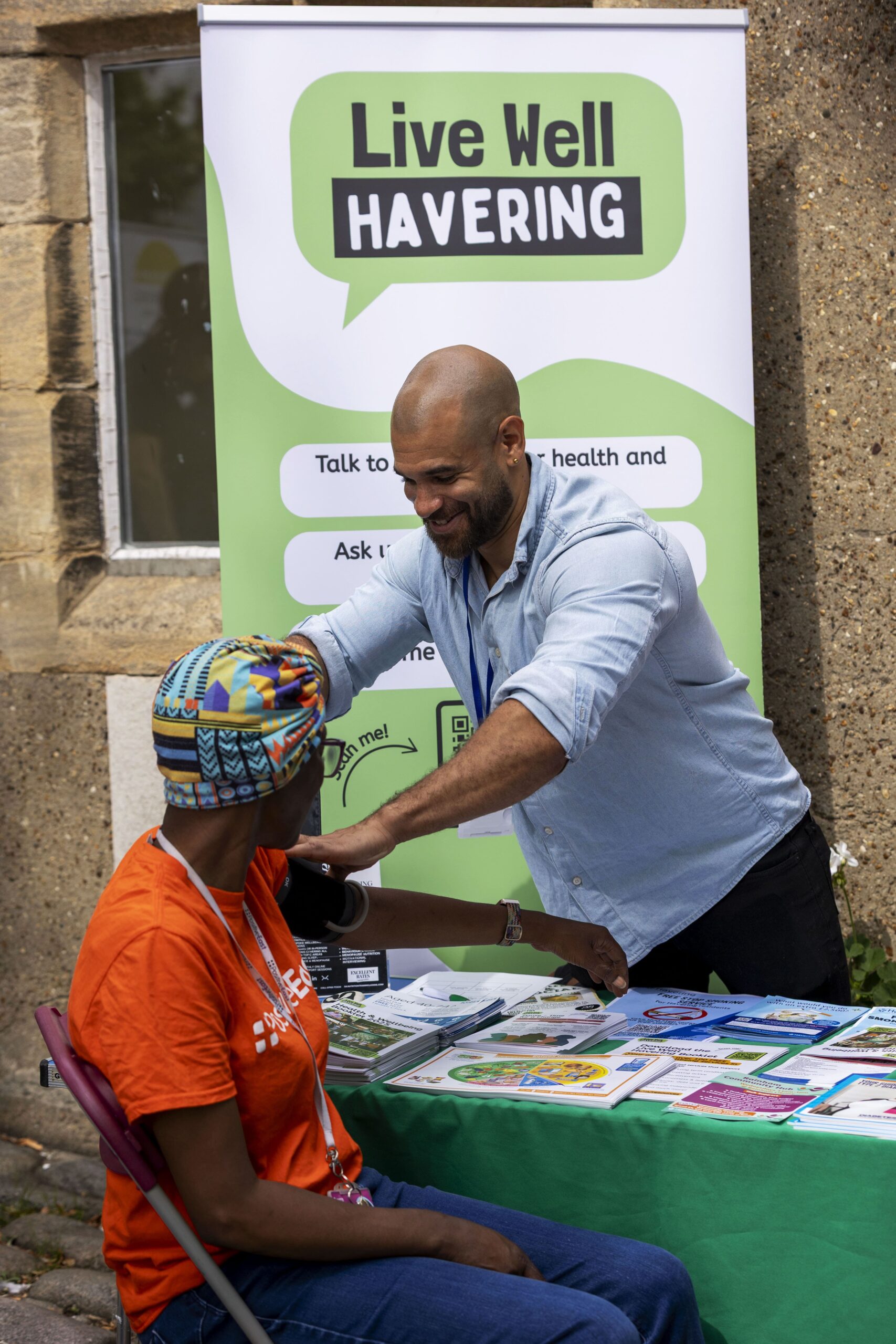 A smiling man in a light blue shirt checks a woman’s blood pressure at an outdoor health event, with a “Live Well Havering” banner in the background. The woman wears an orange shirt and a colorful headwrap.