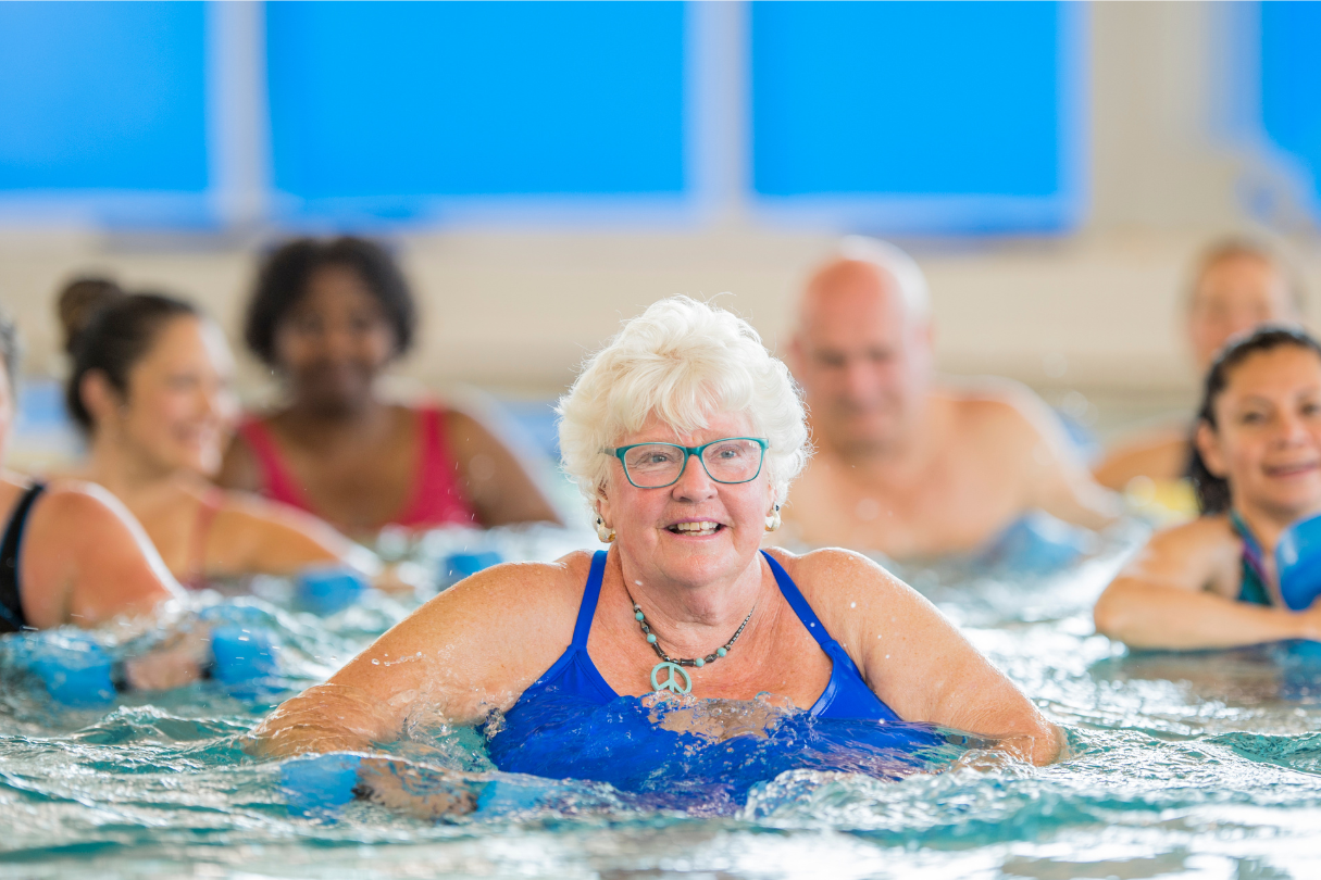 A group of people are engaged in a water exercise class inside an indoor swimming pool. They are using foam dumbbells to perform aquatic workouts
